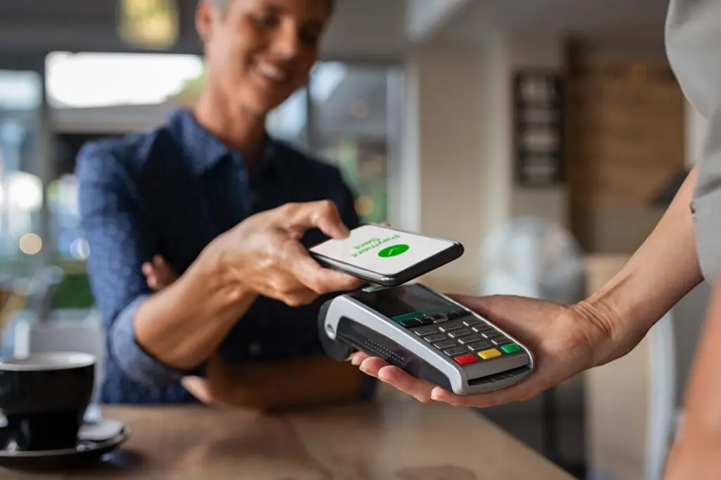 Mature woman paying bill through smartphone using NFC technology in a restaurant. Satisfied customer paying a small business owner through mobile phone using contactless technology. Close up hands of mobile payment at a coffee shop.