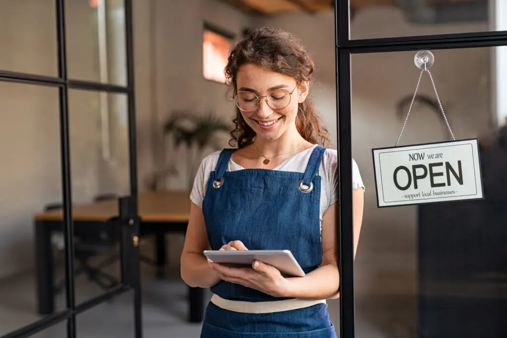 Small businesses in the South utilize Comcast Business to run their organization with fast, secure and reliable Internet and power their mobile devices. IMAGE: Portrait of happy waitress standing at restaurant entrance holding digital tablet to check the reservations. Smiling owner in blue apron standing at coffee shop entrance leaning on door with open signboard. Young woman using working at coffee shop.