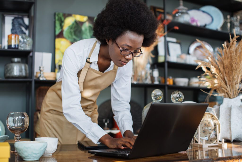 Small Business owners in the South appreciate the power of Comcast Business and its network to keep mobile devices online with fast, secure and reliable internet. IMAGE: Focused afro american woman in eyewear and apron using wireless laptop during working process at shop with various decor. Creating website for online shopping.